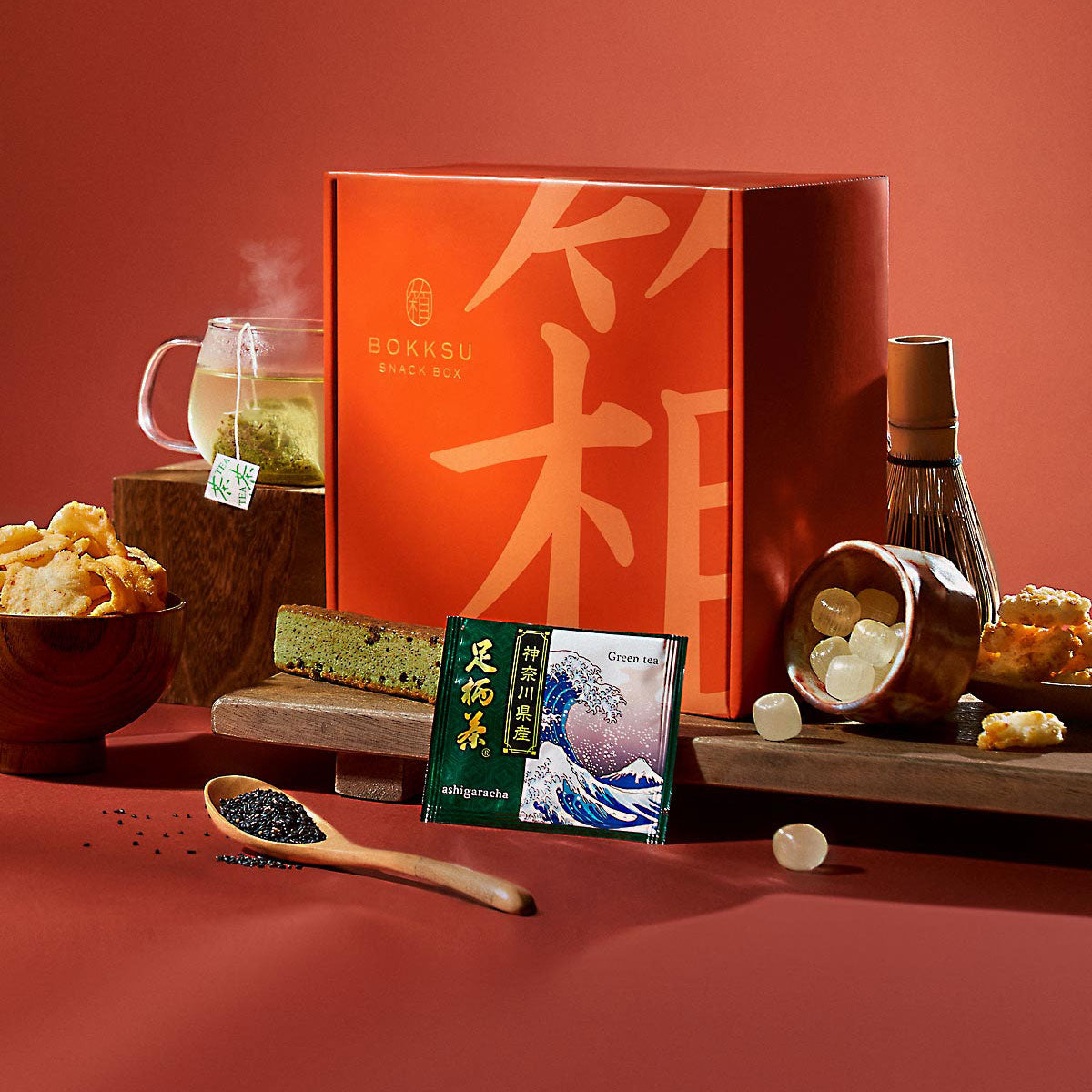 The Bokksu Snack Box: Seasons of Japan includes a variety of Japanese snacks and items like a tea set, crackers, a matcha bar, and candies arranged on a tabletop, with the Bokksu box in the background to celebrate the four seasons.