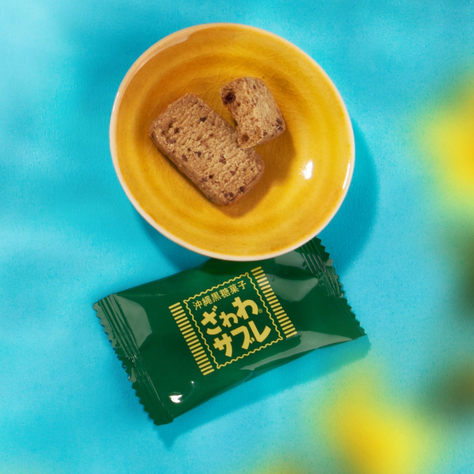 A yellow bowl with two brown biscuits rests above a green Bokksu Snack Box: Seasons of Japan wrapper on a blue and yellow background.
