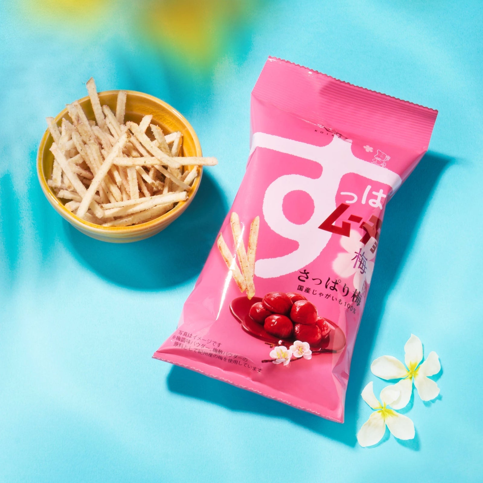 A pink bag from the Bokksu Snack Box: Seasons of Japan sits beside a small yellow bowl of Japanese snack sticks, all displayed on a blue surface with white flowers nearby.