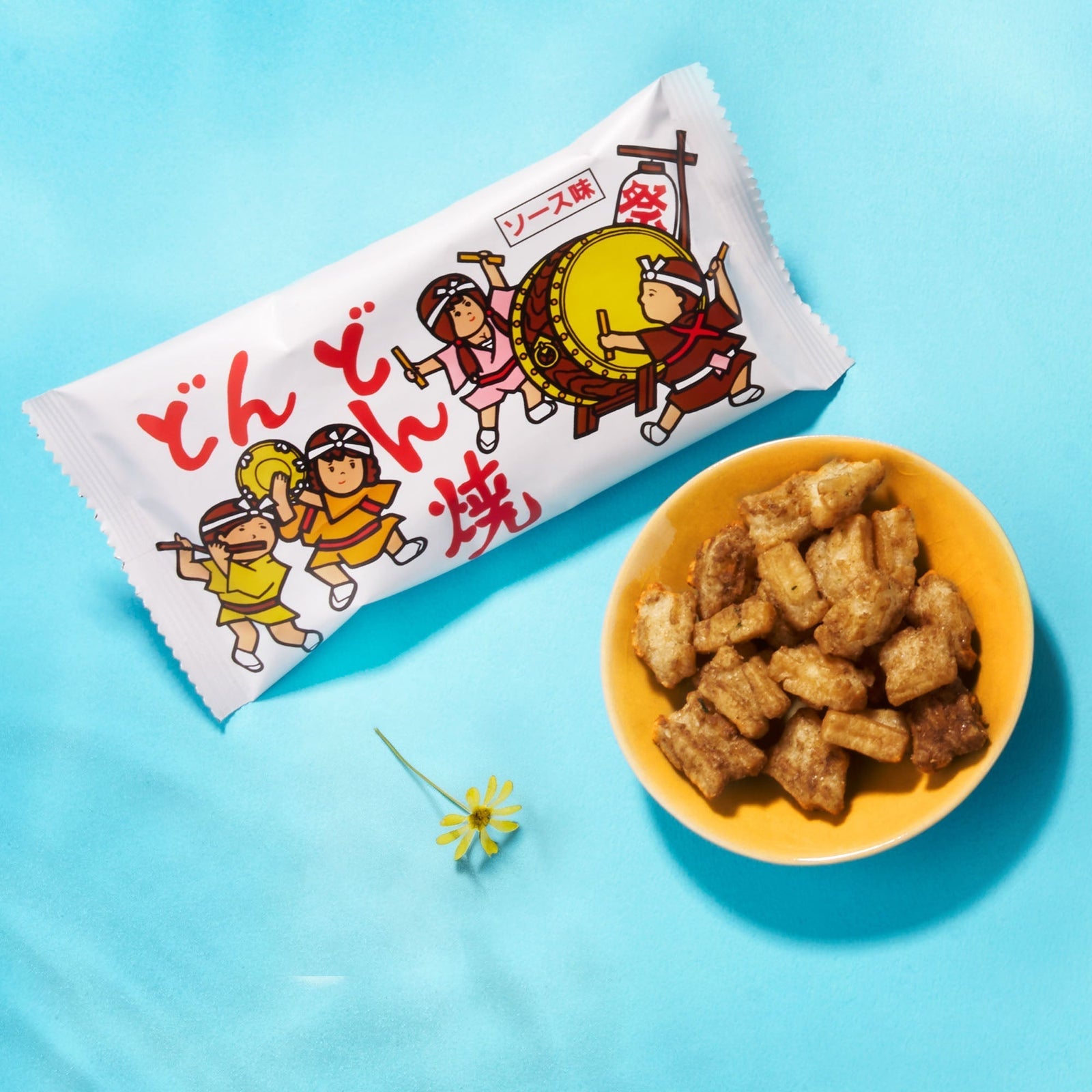 A packaged Japanese snack with cartoon drummers on the wrapper sits by a yellow bowl of brown, bite-sized pieces on a blue surface—an ideal treat to include in your Bokksu Snack Box: Seasons of Japan.