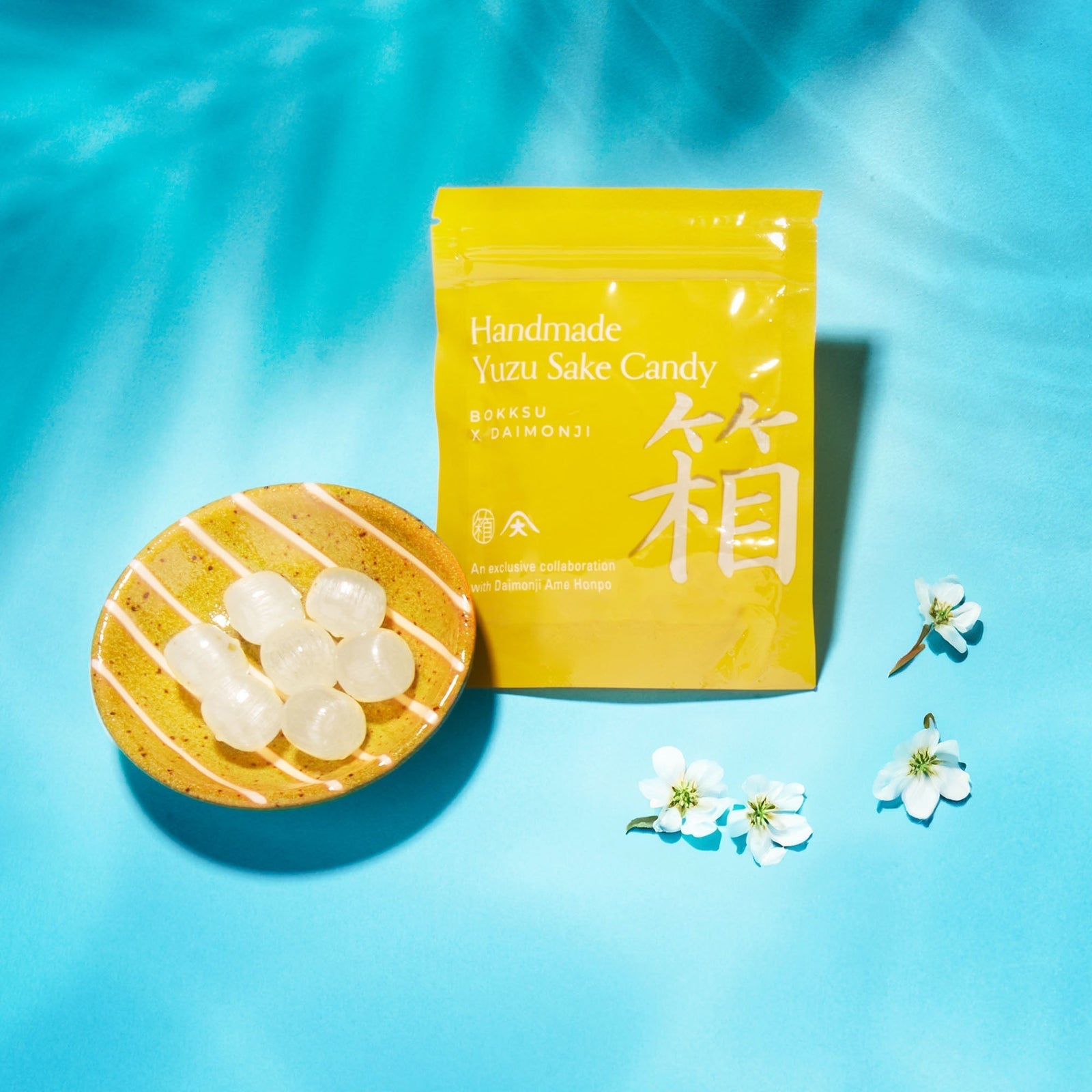A yellow bag labeled "Handmade Yuzu Sake Candy" from the Bokksu Snack Box: Seasons of Japan sits beside a plate of round candies, surrounded by white flowers on a blue background, evoking the charm of unique Japanese snacks.