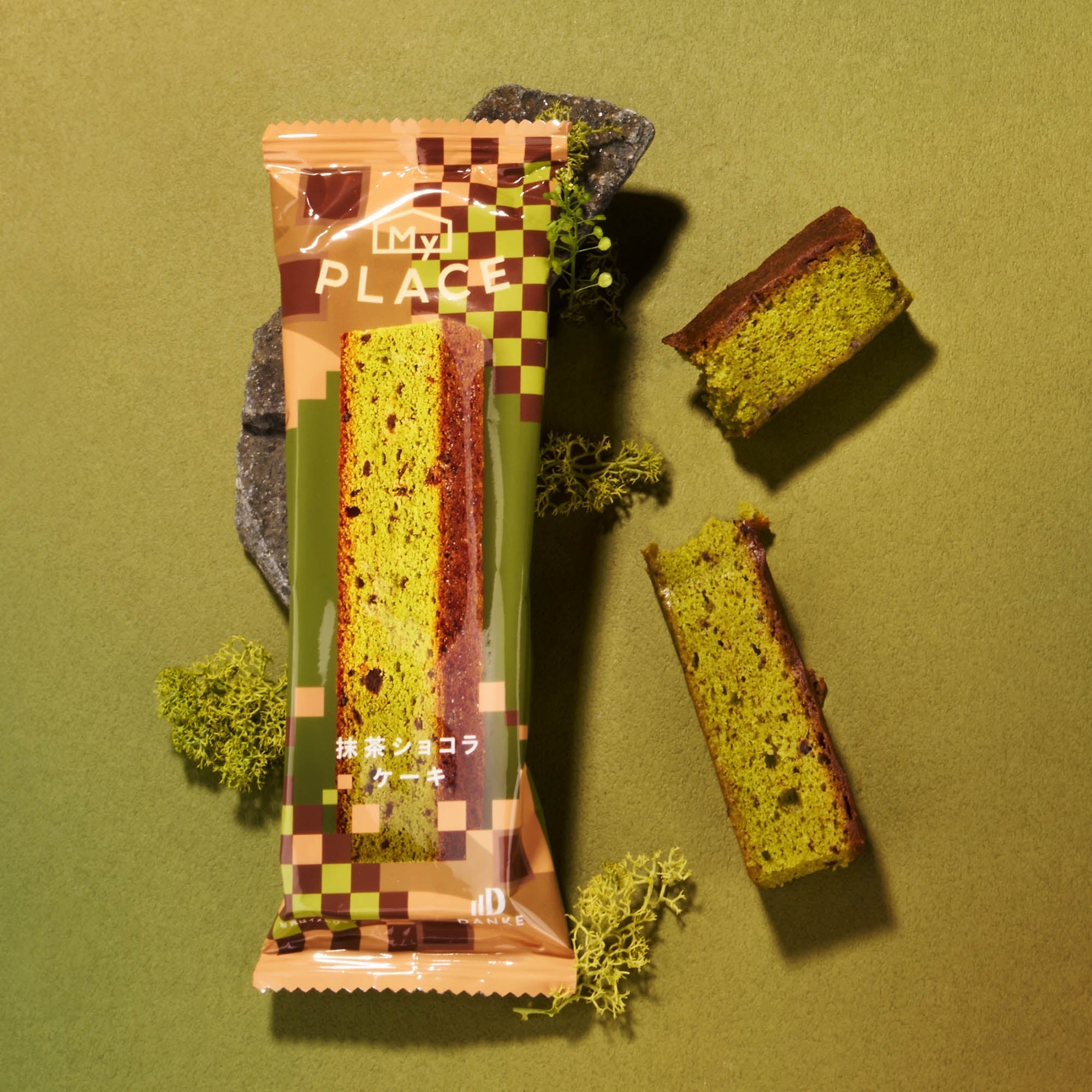 A packaged slice of green and brown matcha cake with unwrapped pieces and moss on a green surface captures the authentic flavors featured in the Bokksu Snack Box: Seasons of Japan.