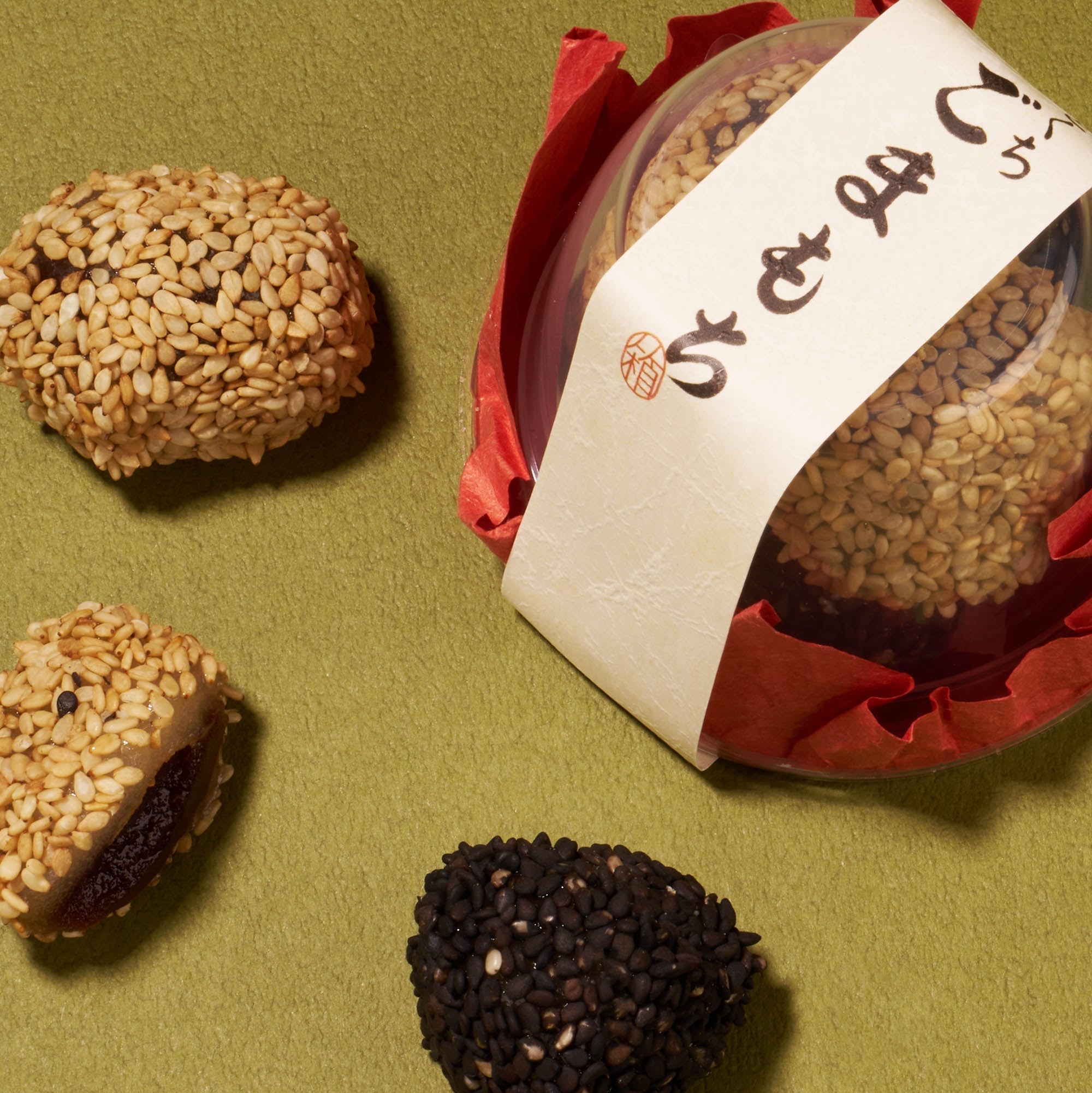 Three sesame-coated Japanese snacks, one in a clear round container with red paper and Japanese writing, rest on an olive green surface—a refined treat from the Bokksu Snack Box: Seasons of Japan.
