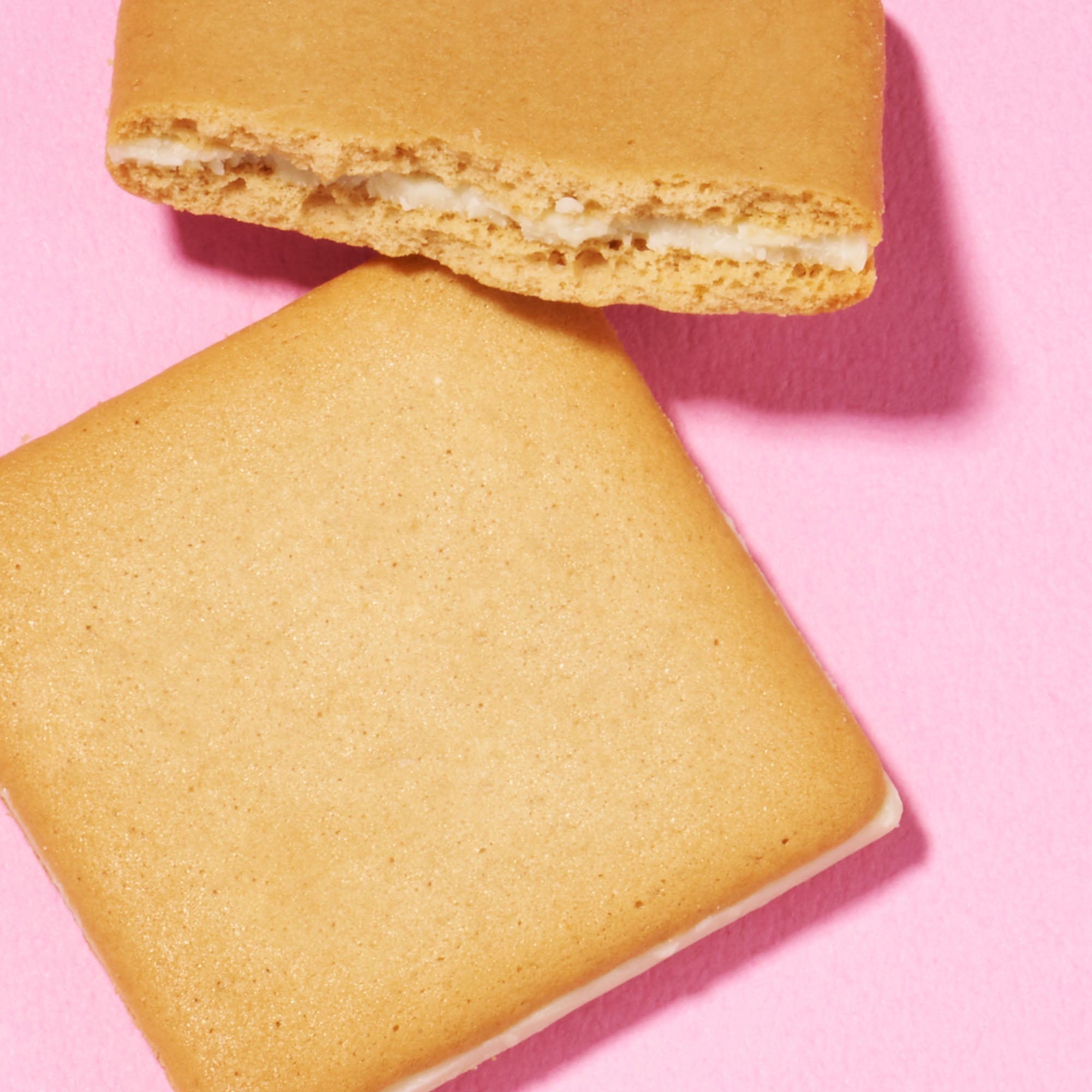 Two square, tan cookies on a pink surface: one whole and one with a bite revealing white filling—an elegant treat featured in the Bokksu Snack Box: Seasons of Japan.
