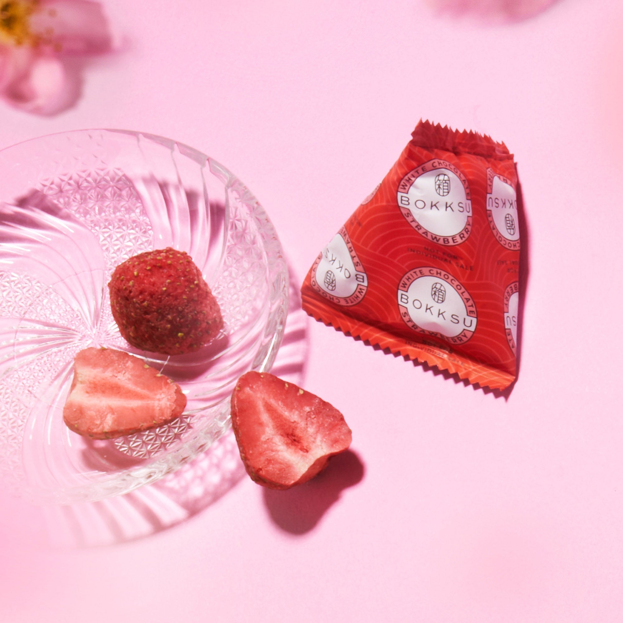 A clear glass dish holds two freeze-dried strawberry pieces and a coated whole strawberry beside a red Bokksu Snack Box: Seasons of Japan packet on a pink surface, highlighting the unique Japanese snacks in every box.