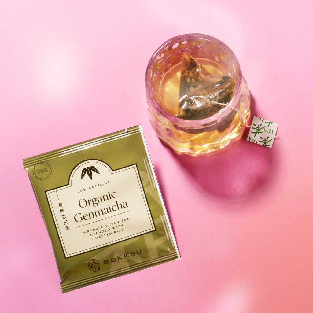 A packet of Bokksu Organic Genmaicha tea and a glass cup of steeping tea on a pink surface—an ideal companion to your Bokksu Snack Box: Seasons of Japan.