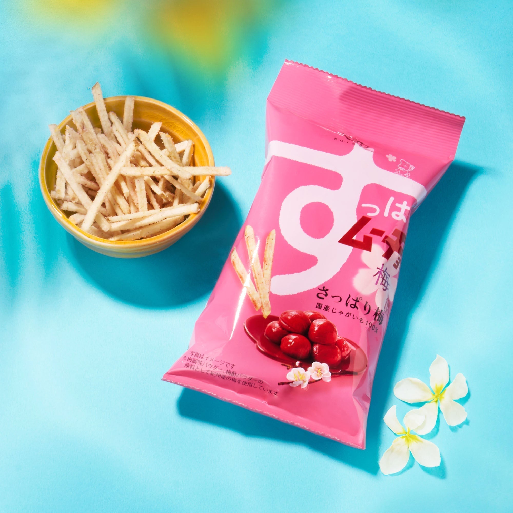 A pink bag from the Bokksu Snack Box: Seasons of Japan sits beside a small yellow bowl of Japanese snack sticks, all displayed on a blue surface with white flowers nearby.